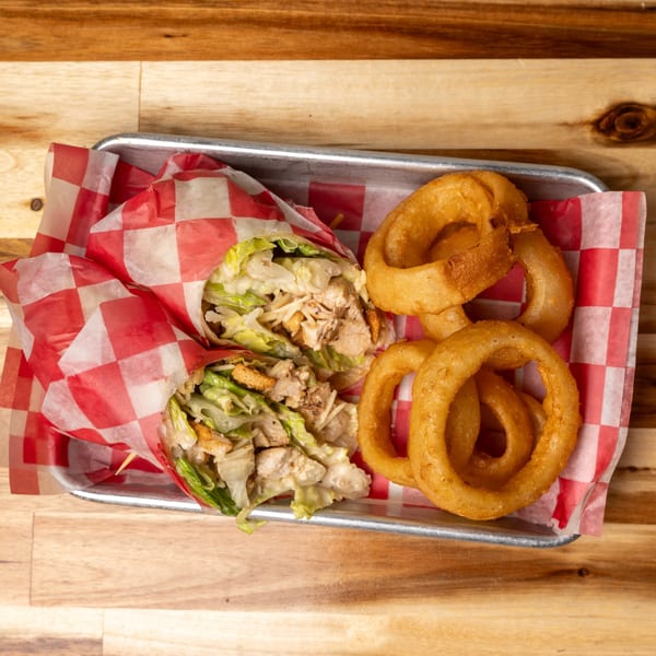 Two chicken Caesar wraps and a side of golden onion rings on a metal tray with red checkered paper.