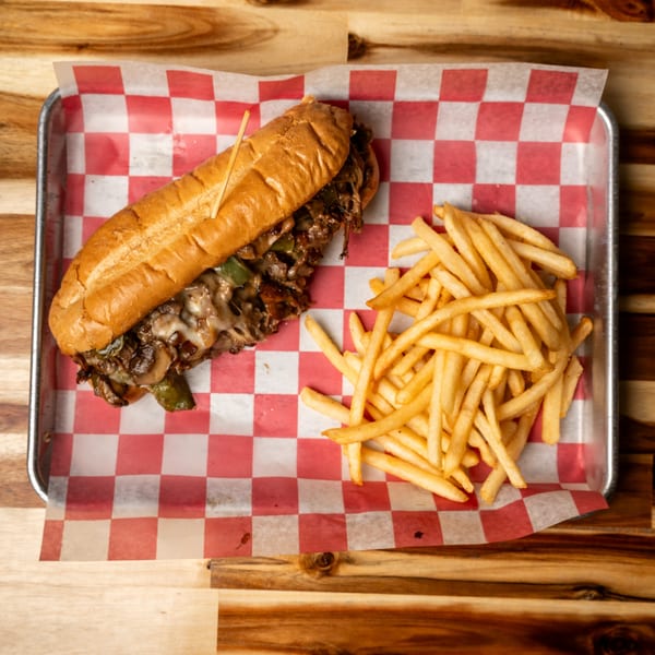 A Jaxx Wing Co. cheesesteak sandwich and a pile of fries on a metal tray with checkered paper.